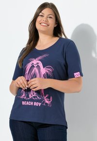 Usmívající se žena v námořnickém modrém tričku s grafikou růžového psa a kraba a textem "BEACH BOY", v kombinaci s tmavými džíny.