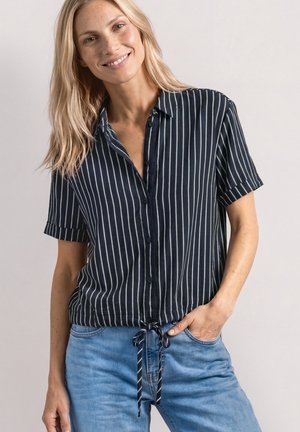 Uśmiechnięta kobieta z długimi blond włosami, ubrana w granatową koszulę z krótkim rękawem, białe pionowe paski i jasnoniebieskie dżinsy, z jedną ręką w kieszeni.
