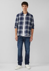 Blauw-wit geruit flanellen overhemd met lange mouwen en knopen aan de voorkant, gedragen over een wit T-shirt, gecombineerd met donkere spijkerbroek en witte sneakers.