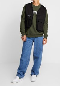 Chaleco negro con múltiples bolsillos sobre una sudadera verde, combinado con vaqueros azules holgados y zapatillas blancas, mostrando un look casual en capas.