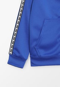 Sudadera azul con bolsillo canguro, puños acanalados y paneles laterales con la marca "NIKE" en blanco y negro repetida. Textura de tela suave.