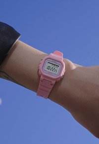 Montre numérique rose avec un cadran carré et un écran clair, dotée d'un bracelet en plastique. La montre indique l'heure comme étant 10:22:25.