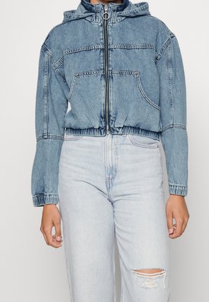 Lichtblauwe cropped spijkerjas met ritssluiting, elastische taille en manchetten, gedragen met high-waisted lichtblauwe gescheurde jeans.