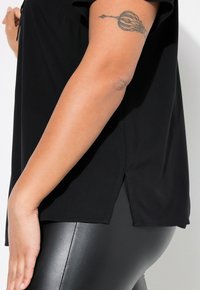 Top nero realizzato in morbido tessuto con maniche corte e spacchi laterali, abbinato a leggings neri. Tatuaggio visibile sul braccio sinistro.