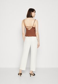 Top marrone senza maniche con spalline sottili, caratterizzato da un design fluido e una schiena con doppie spalline. Abbinato a pantaloni bianchi a gamba larga e tacchi neri.