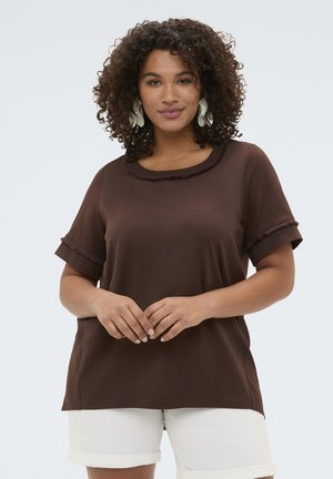 Krøllet kvinde iført en brun kortærmet top med tekstureret kant og hvide shorts, står med hænderne foldet, neutralt baggrund.