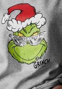 Sweat-shirt gris avec un graphique du Grinch portant un chapeau de Père Noël rouge et des lunettes de soleil argentées scintillantes. Le texte "The Grinch" est inscrit sous l'image.