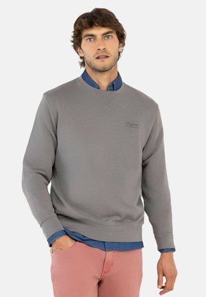 Hombre con sudadera gris sobre camisa azul y pantalones rosas, de pie con una mano en el bolsillo sobre fondo blanco.