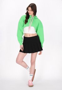 Felpa verde corta con lacci rosa, abbinata a una gonna plissettata nera e sneakers rosa. Calzini bianchi con righe verdi.