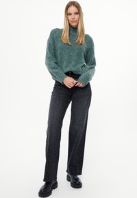 Groene gebreide trui met een hoge halslijn, gecombineerd met zwarte wijde broek met kleine, zilveren studs en chunky zwarte laarzen.