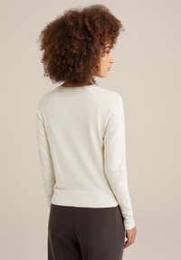 Gaišas krēma krāsas vilnas džemperis ar mīkstu tekstūru, ribināta apakšmalu un piedurknēm, garām piedurknēm, apaļu kakla izgriezumu, vienkāršs dizains, valkāts kopā ar tumšiem biksēm.