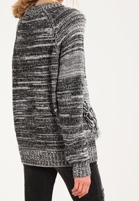 Osoba ubrana w czarno-białą sweter z melanżem, ozdobiony frędzlami na rękawach, w komplecie z czarnymi spodniami, stojąca na tle białym.