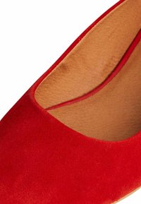 Scarpa rossa in suede con texture liscia, punta arrotondata e rivestimento interno color beige. I bordi superiori presentano una curva sottile.