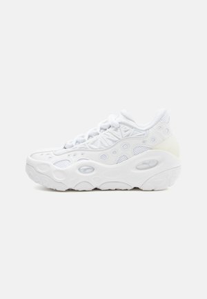 Vit sportig sneaker med en strukturerad mesh-övre, rund tå och en kraftig sula med cirkulära utskärningar för ventilation.