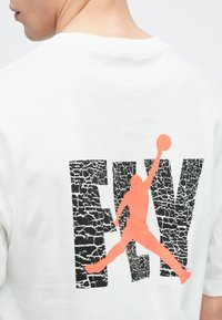 Λευκό μπλουζάκι με μεγάλο μαύρο ραγισμένο κείμενο "FLY" και πορτοκαλί σιλουέτα παίκτη που πηδά για να σουτάρει ένα μπάσκετ στην πλάτη.