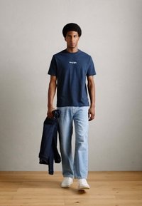 Camiseta de algodón azul marino con el texto blanco "Fuera de la oficina", combinada con jeans azul claro y zapatillas blancas, sosteniendo una chaqueta de mezclilla.