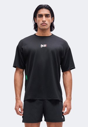 Mand med mørkt hår iført en ensfarvet sort t-shirt og sorte shorts, vendt fremad mod en ensfarvet lys baggrund.