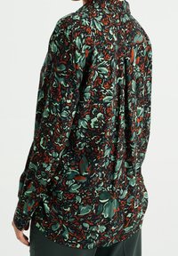 Mörkgrön långärmad skjorta med blommönster i rost och teal, bärs av person som står med ryggen mot kameran framför en vit bakgrund.