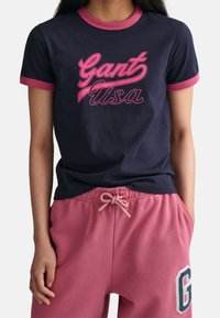 Camiseta de marina con cuello rosa, con "Gant USA" en cursiva. Combinada con pantalones de chándal rosas, que muestran un cordón de ajuste y un logo bordado.