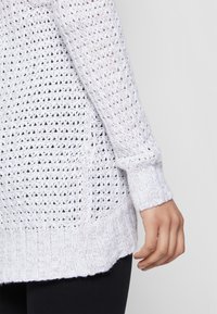 Pull en laine blanc avec un motif en mailles ouvertes, des poignets côtelés et un ourlet texturé. Le design est léger et légèrement oversized.