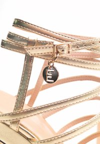 Sandalo con cinturini in oro metallizzato con una superficie liscia, dotato di un charm logo nero e argento. Cinture multiple si incrociano sopra la soletta.