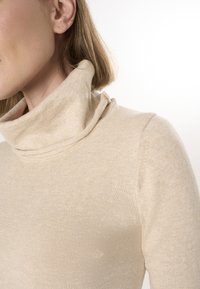 Beige turtleneck sweater i blødt, fint strikket materiale med en foldekrave og tætsiddende pasform. Nærbillede fanger tekstur og sømdetaljer.
