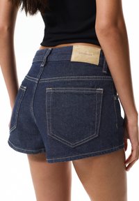 Tmavé denim šortky so priliehavým strihom, s koženou etikettou na páse a kontrastným šitím. Zadné vrecká s zakriveným tvarom.