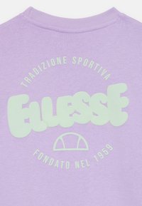 Levandulová mikina se velkým, světlezeleným logem "Ellesse", s textem "Tradizione Sportiva" a "Fondato nel 1959" nahoře a dole.