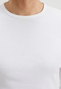 Camiseta de algodón blanca con un cuello redondo ribeteado. Textura suave, forma ajustada y mangas largas, sin patrones ni acentos visibles.