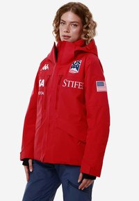 Giacca impermeabile rossa con cappuccio, dotata di numerose tasche con zip, logo della squadra olimpica sciistica degli Stati Uniti, toppa della bandiera americana e polsini regolabili.