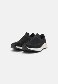 Des chaussures noires à enfiler avec une tige en tricot respirant, surface texturée et semelle en caoutchouc blanc avec une subtile accent tan au niveau du talon.
