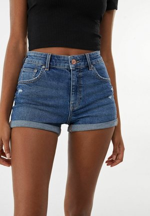 Kvinde iført højtaljede blå denimshorts med opsmøg og en sort cropped kortærmet top mod en ensartet lys baggrund.