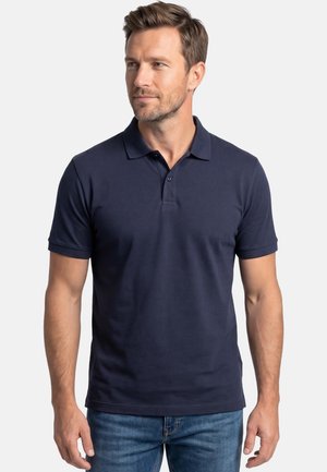 Mand iført en mørkeblå kortærmet poloshirt og blå jeans, kigger let mod højre mod en hvid baggrund.