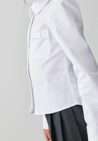 Bambino che indossa una maglietta bianca a maniche lunghe con un fiocco sulla tasca e una gonna grigia plissettata su uno sfondo semplice.