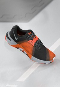 Black and bright orange athletic shoe with gray laces, Nike logo, and patterned sole resting at an angle on a gray surface.