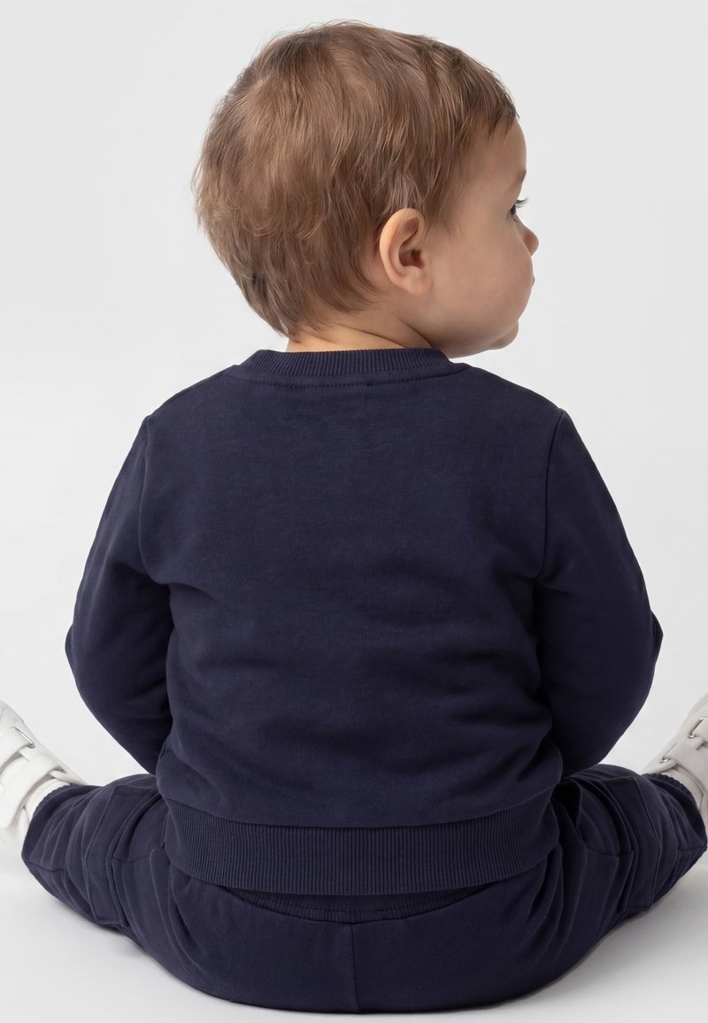 Tout-petit aux cheveux châtain clair assis par terre de côté, portant une tenue bleu marine et des chaussures blanches.