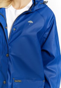 Blå vattentät jacka i slätt tyg, med en silverfärgad paraplylogotyp, dragkedjestängning och tryckknappar framtill.