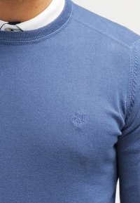 Blauer Strickpullover mit rundem Halsausschnitt und gerippten Details. Hat eine strukturierte Oberfläche und ein kleines, auf der Brust gesticktes Logo.