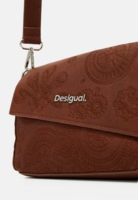 Borsa a tracolla in pelle marrone con embossing, caratterizzata da motivi floreali e un logo in metallo. Ha una chiusura a patta e una tracolla regolabile con clip.