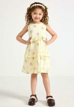 Pige med krøllet hår iført en gul blomstret kjole med flæser, margueritpandebånd og mørkebrune fåreskindskloger står med hænderne på hofterne.