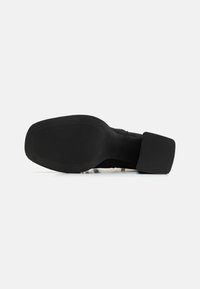 Semelle de chaussure noire avec une semelle en caoutchouc texturée, un design incurvé et un talon plat. Le matériau semble robuste et durable.