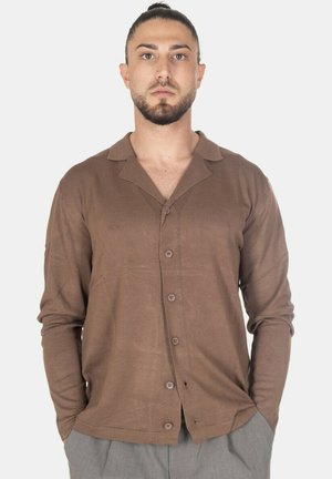 Uomo con capelli scuri raccolti in uno chignon, indossa un cardigan marrone con bottoni e pantaloni grigi, in piedi con le mani in tasca davanti a uno sfondo uniforme.