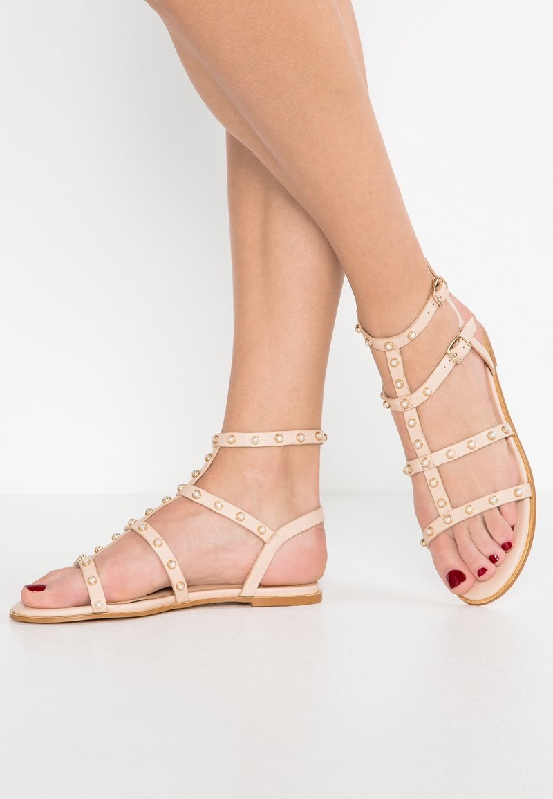 Pieds portant des sandales gladiateur plates beige avec des clous en perle et des brides à la cheville sur fond blanc.