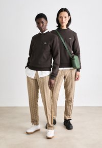 To personer har på seg brune hettegensere med et logo, hvite skjorter under, og mønstrede beige bukser med sidestriper, kombinert med joggesko.