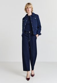 Persona bionda che indossa un giacca trapuntata blu navy, jeans a gamba larga scuri, maglia blu navy e décolleté borgogna a punta, in piedi contro uno sfondo neutro.
