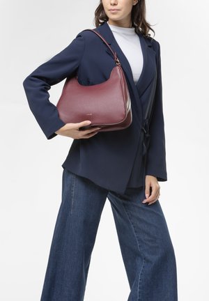 borsa a tracolla in pelle bordeaux con texture liscia, forma curva e una sola tracolla, tenuta da una persona in blazer blu marina e jeans a gamba larga.