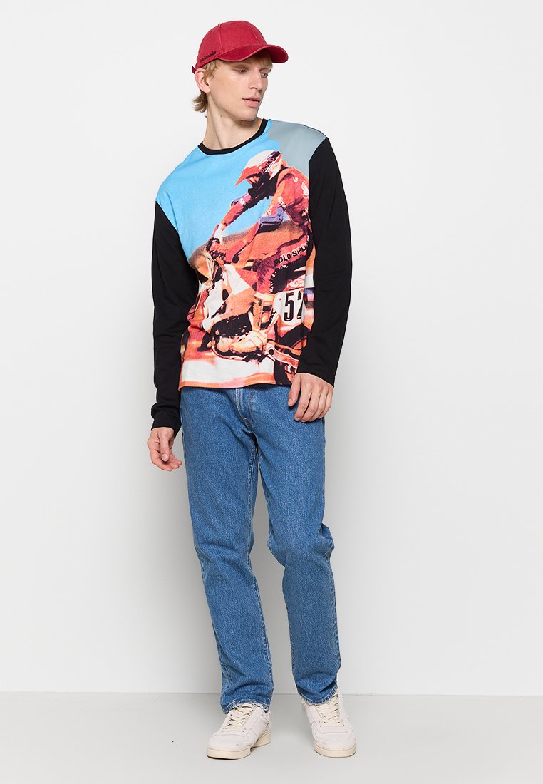 Langärmliges Hemd mit einem Grafikdesign in Blau und Orange mit Motorradmotiv, kombiniert mit blauen Jeans und weißen Turnschuhen.