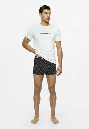 Hombre con camiseta blanca de Karl Lagerfeld y bóxers negros a cuadros, de pie descalzo sobre un fondo blanco liso.