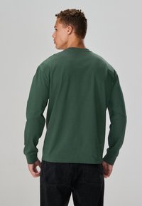 Long-sleeve green cotton shirt with a crew neck, featuring a smooth texture and straight hem, paired with dark jeans.