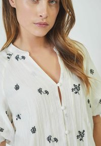 Camicia in tessuto leggero bianco con ricami floreali neri, scollo a V e chiusura con tre bottoni. Presenta maniche corte e una finitura testurizzata.